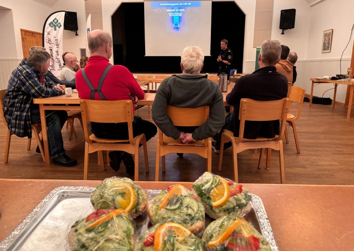 Informationsmöte i Vinbergs bygdegård om GPS-stölder, med deltagare som lyssnar på polisens presentation och en bricka med smörgåsar i förgrunden.