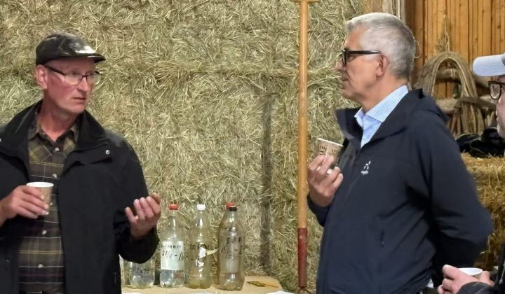 Två personer samtalar framför en vägg av höbalar. Den ena håller en pappersmugg, den andra ett mindre föremål. På bordet framför dem står plastflaskor med etiketter och olika vätskor. I bakgrunden syns verktyg och en träpinne som lutar mot höbalen.