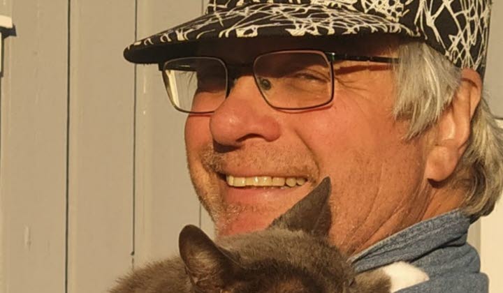En person med keps, glasögon och grått hår ler och håller en grå katt med vita tassar på axeln. Katten vilar med ögonen stängda. Bakgrunden är en ljusgrå trävägg.