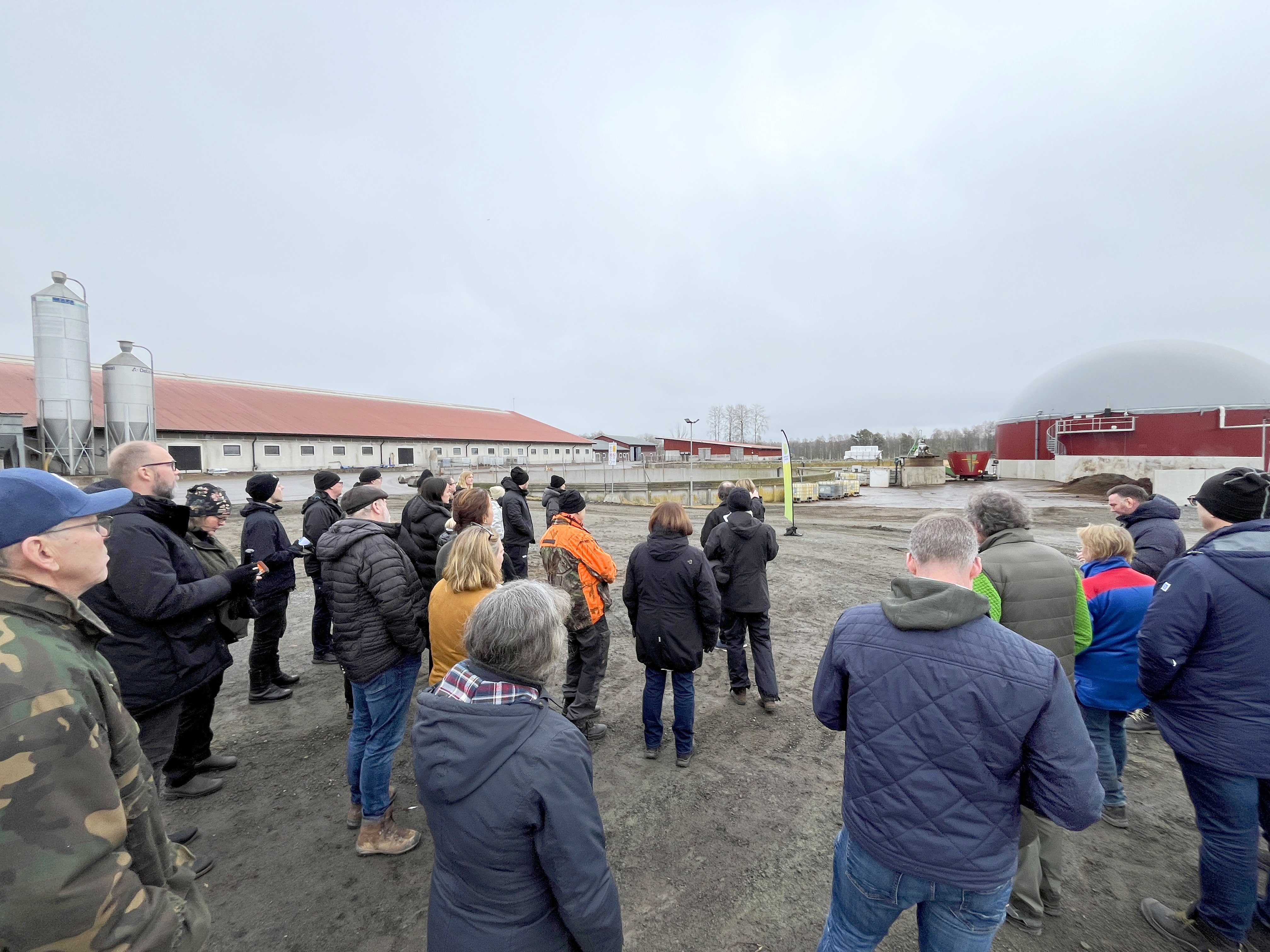 En samling personer står på en gårdsplan på en mjölkgård och tittar på en biogasanläggning.