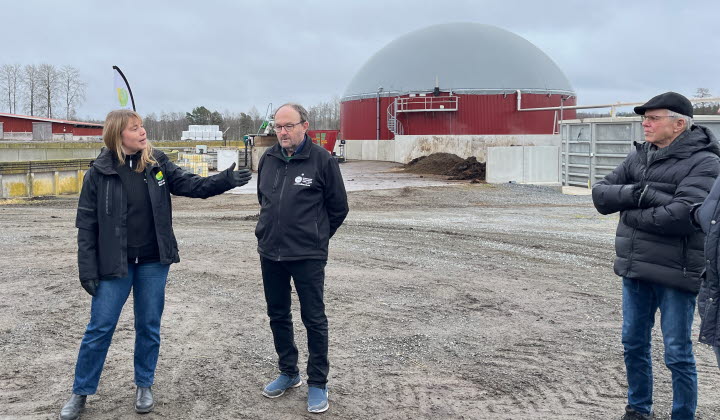 Tre personer står på en gårdsplan framför en rötanläggning för biogas. 