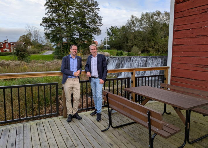 Två män står i blåa kavajer står avslappnat lutade mot ett altanräcke.