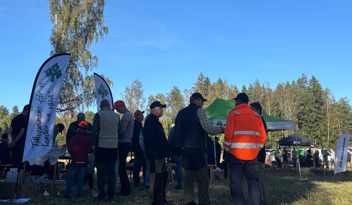 En grupp människor står samlade utomhus på en gräsbevuxen yta med skog i bakgrunden. Flera tält och beachflaggor syns. En person bär en orange jacka. Det är dagsljus och klart väder.