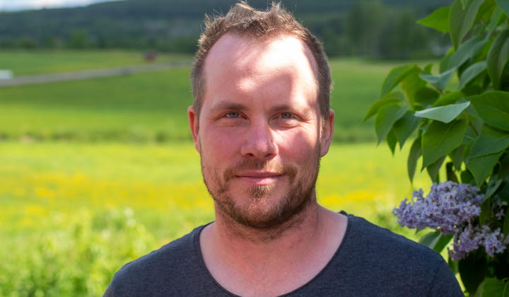 En man tittar in i kameran, i bakgrunden syns syrenen blomma samt vidsträckta ängar och marker.