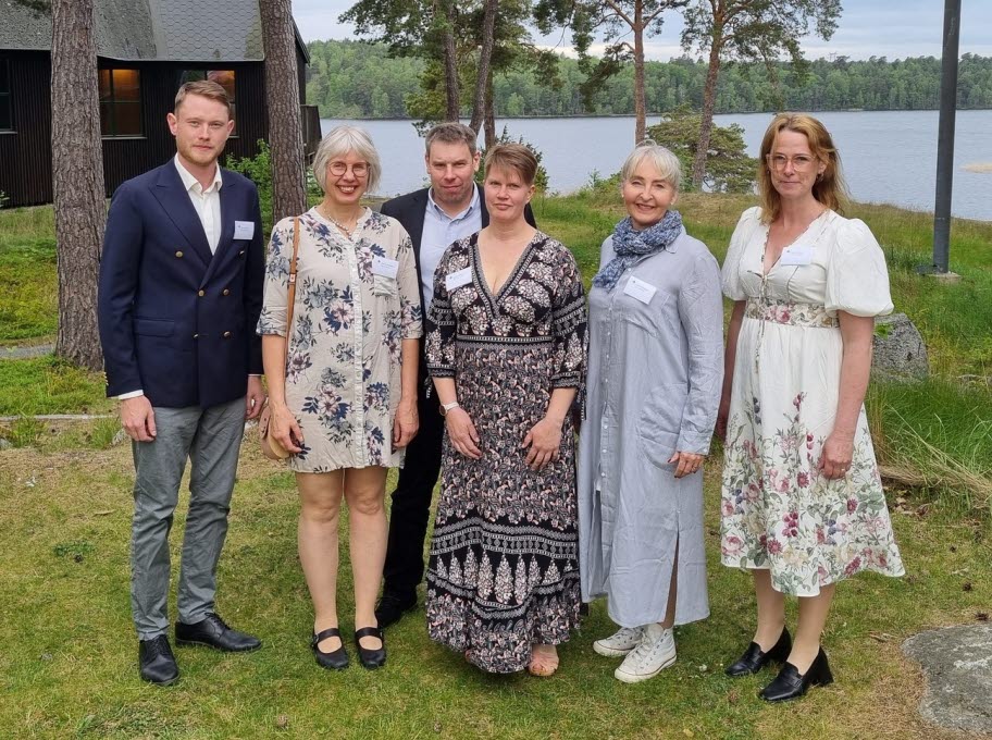 Sex personer står finklädda framför hus, tallar och en sjö. 
