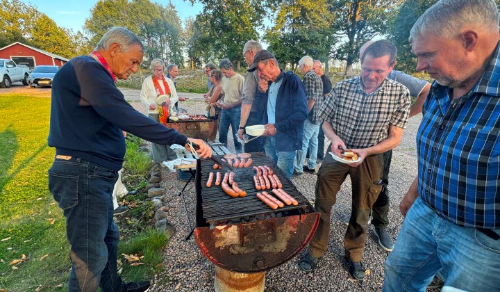 män och kvinnor står utomhus kring en grill, fyld med grillkorv